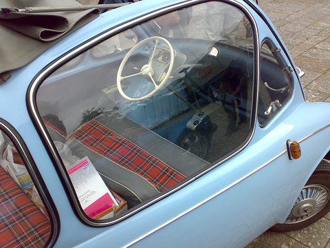 Tartan bench and white steering wheel seen through the side window of a pale blue car. Opens a larger view