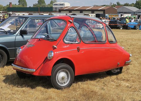 Red bubble car on dry grass seen from the front left, an old saloon and hangars behind. Opens a larger view