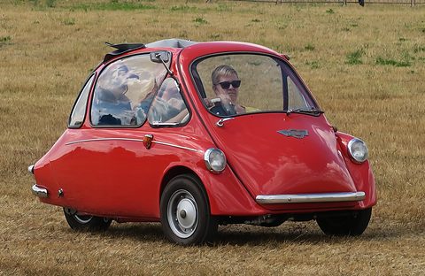 Red bubble car with its roof open driving on dry grass, the driver in sunglasses. Opens a larger view