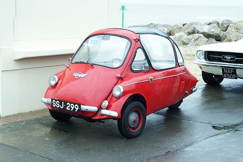 Red bubble car parked on a wet promenade with the sea and rocks behind. Opens a larger view
