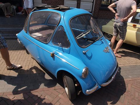 Blue bubble car on brick paving with its roof rolled back, legs of passers-by at the edge. Opens a larger view
