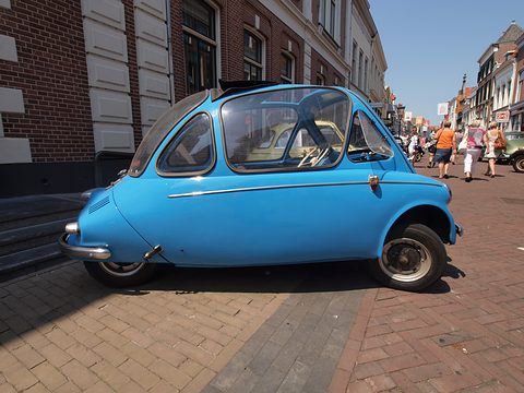 Blue bubble car in profile on brick paving, shoppers in the street behind. Opens a larger view
