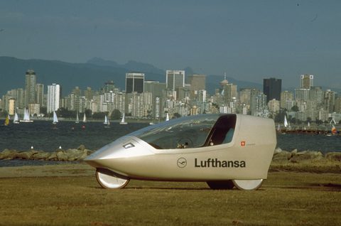 A long low silver pod-shaped vehicle with a clear canopy and Lufthansa lettering on grass by the sea, skyscrapers beyond.