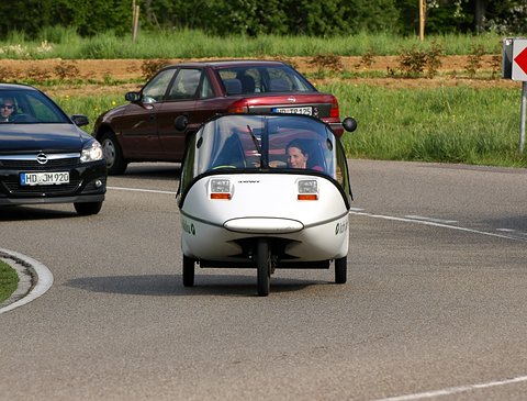 White three-wheeler with a clear canopy and its driver visible cornering on a road, cars and fields behind. Opens a larger view