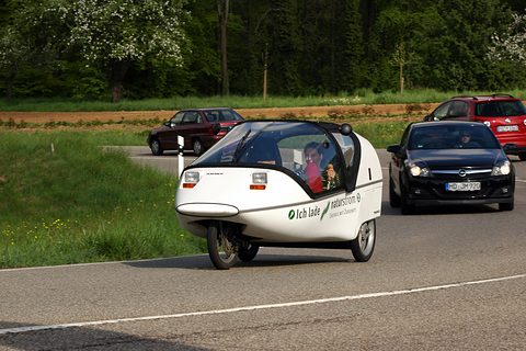 Side view of a white three-wheeler with two occupants on a road, a black hatchback and green fields behind. Opens a larger view