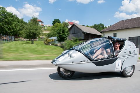 Silver three-wheeler with an open canopy driving past farm buildings and a green slope under blue sky. Opens a larger view