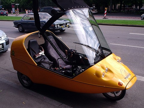Orange three-wheeler with a clear screen and hood lifted, showing two black seats, parked by a road. Opens a larger view