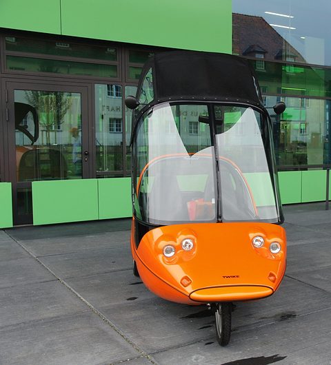 Front of an orange three-wheeler with four small round lamps and a tall tipped-up hood, on concrete paving. Opens a larger view