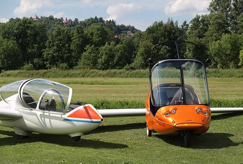 Front of an orange three-wheeler with a tall screen beside the long white wing of a glider on grass. Opens a larger view