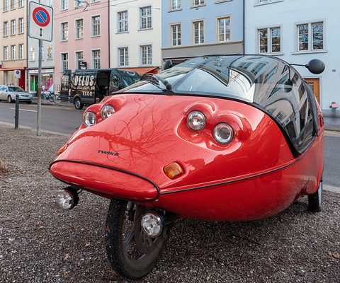 Front of a red three-wheeler with four round lamps and a dark canopy on gravel, town houses behind. Opens a larger view