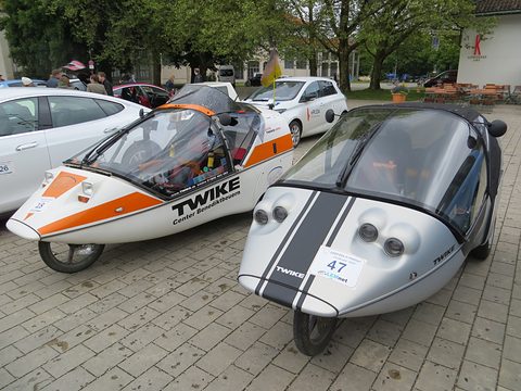 Two silver and white three-wheelers with tinted canopies, one with orange stripes, parked on paving with start numbers. Opens a larger view