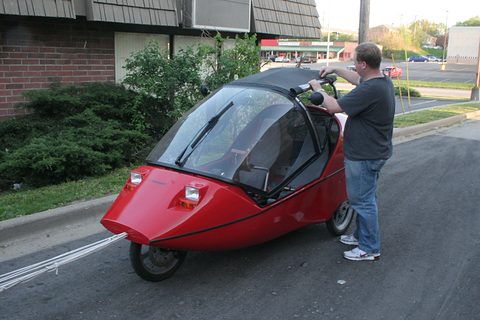 Red three-wheeler with a tinted canopy tipped open, a man in a grey T-shirt bending over it at the kerb. Opens a larger view