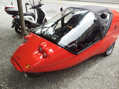 Red three-wheeler with a dark tinted canopy and four round nose lamps on gravel beside a tree and a scooter. Opens a larger view