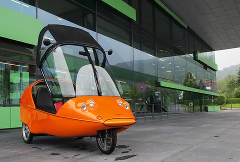 Orange three-wheeler with twin round lamps and a black hood tipped up, on concrete in front of green glazing. Opens a larger view