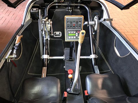 Two black leather seats either side of a central joystick, switch panel and pedal crank on a dark carpet. Opens a larger view