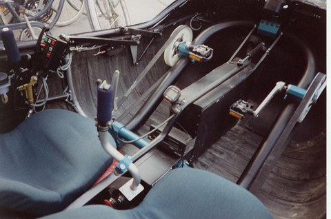 Pedal crank with two pedals on a chain housing in a carpeted footwell, a joystick and blue seats either side. Opens a larger view