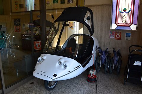 White three-wheeler with a black hood and four round lamps on a speckled floor, wheelchairs lined up behind. Opens a larger view