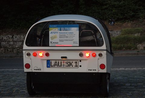 Rear of a white three-wheeler with rows of round red lamps, two rear wheels and a solar-power advert in the window. Opens a larger view