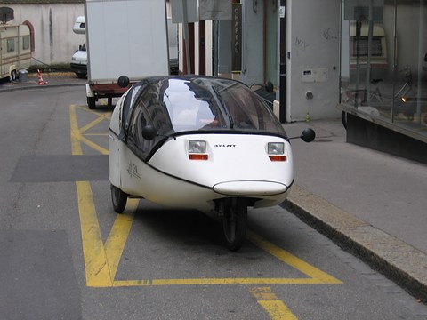 White three-wheeler with a dark canopy and twin lamps standing on yellow road markings beside a shop front. Opens a larger view