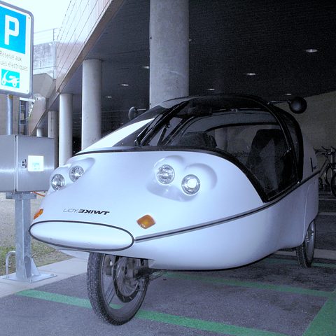 Front of a silver three-wheeler with twin round lamps either side of the nose and a single front wheel. Opens a larger view
