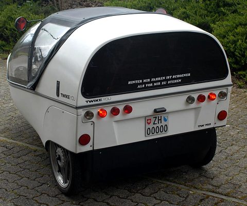 Rear of a white three-wheeler with a dark rear window, round tail lamps and twin rear wheels on block paving. Opens a larger view