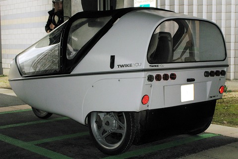 Silver three-wheeler with a long glazed canopy, row of round tail lamps and blanked number plate in a parking bay. Opens a larger view