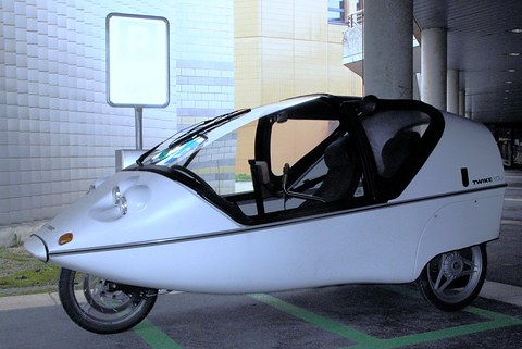 Side view of a silver three-wheeler with an open-framed canopy and three spoked wheels under a concrete deck. Opens a larger view