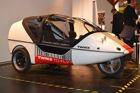 White three-wheeler with a barcode-style red and black livery on a dark show floor, visitors behind. Opens a larger view