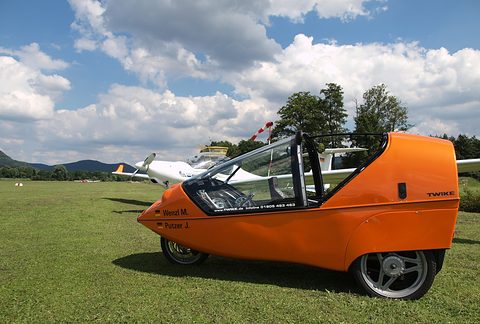 Side view of an orange three-wheeler with an open canopy frame on mown grass, a white glider and clouds behind. Opens a larger view