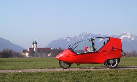 Red three-wheeled two-seater with an open-sided canopy frame on grass, a white abbey and snowy mountains behind. Opens a larger view
