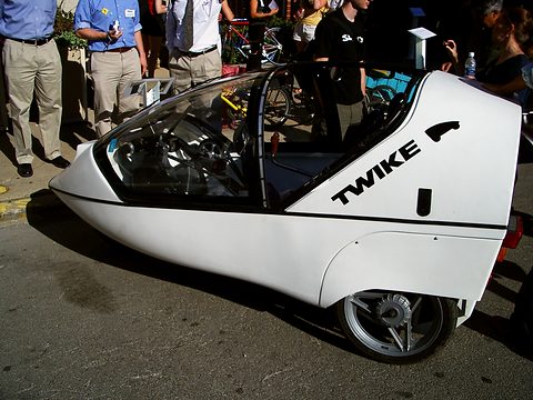 White three-wheeler lettered TWIKE with a raised canopy frame and spoked rear wheel, visitors standing behind. Opens a larger view