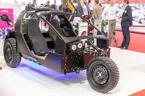Black tubular-framed chassis with two seats, round lamps and chunky tyres, visitors behind. Opens a larger view