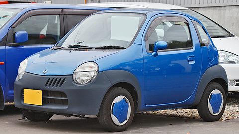 Blue tiny two-seat hatchback with a grey lower body and blue wheel covers parked on gravel. Opens a larger view