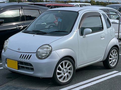 Silver tiny hatchback with multi-spoke alloy wheels and window visors parked between larger cars. Opens a larger view