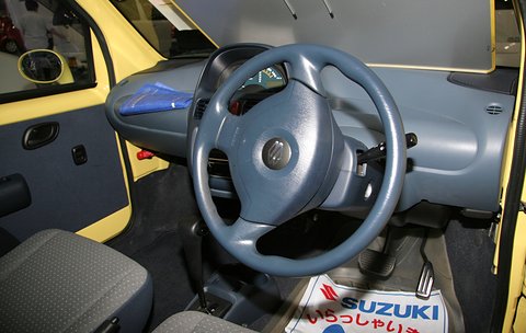 Grey two-spoke steering wheel, a blue-grey dashboard and yellow door trim, with a Suzuki floor mat.