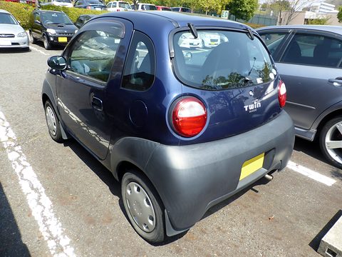 Rear of a dark blue tiny hatchback with round tail lamps and a grey lower body, parked in a marked bay. Opens a larger view