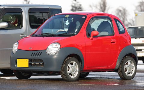 Red tiny two-seat hatchback with a grey bumper and steel wheels, parked beside a grey van. Opens a larger view