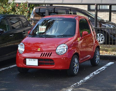 Red tiny two-seat hatchback with a colour-matched lower body in a marked bay beside a black car. Opens a larger view