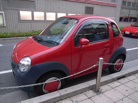 Red tiny two-seat hatchback with a grey lower body parked behind a chain barrier on a city street. Opens a larger view