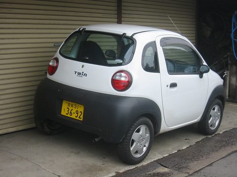 Rear of a white tiny hatchback with round tail lamps, a yellow kei plate and a grey lower body, by a roller shutter. Opens a larger view