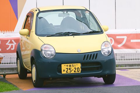 Front of a tiny cream-yellow hatchback with round headlamps, a slotted grille and a yellow kei plate. Opens a larger view