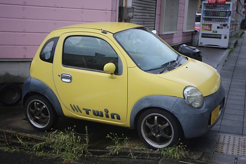 Yellow tiny two-seat hatchback with a grey lower body and twin lettering, parked by a pink building and a vending machine. Opens a larger view