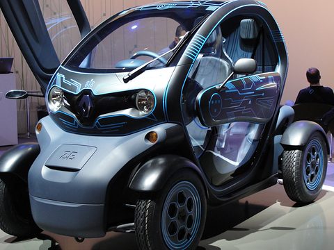 Grey tandem two-seater with glowing blue line graphics and its scissor door raised, a man looking on. Opens a larger view