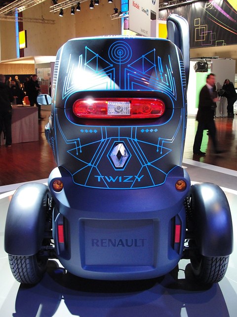 Rear of a dark grey narrow car with a red tail lamp and blue line graphics, visitors at the edge. Opens a larger view