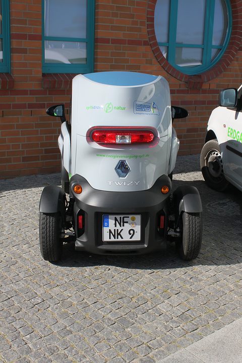 Rear of a narrow white and grey car with a red tail lamp and a German plate, on cobbles by a brick building. Opens a larger view
