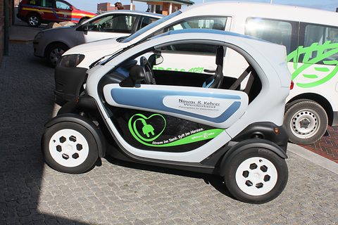 Side view of a white tandem two-seater with company lettering and a green plug graphic, parked by a white van. Opens a larger view