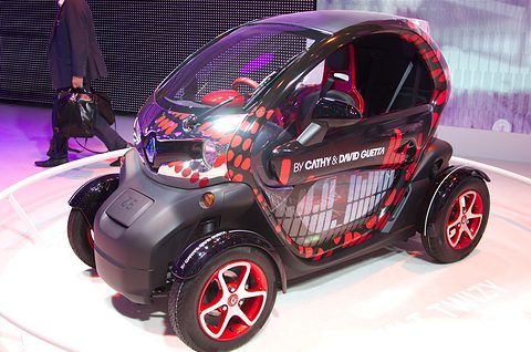 Black tandem two-seater with red dot graphics and red wheels on a white turntable under pink light. Opens a larger view