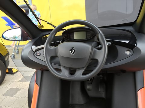 Steering wheel with a Renault badge, a small digital display and pedals, seen from the driver’s seat through the screen. Opens a larger view