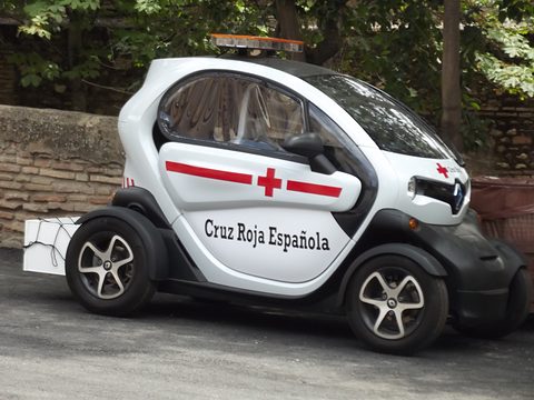 White tandem two-seater marked Cruz Roja Española with a roof light and a box on its tail, parked by a wall. Opens a larger view