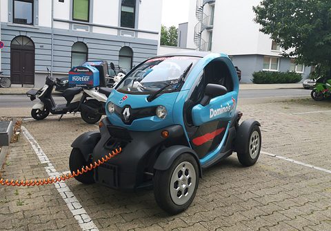 Blue and black tandem two-seater with Domino’s lettering connected to an orange charging cable, scooters behind. Opens a larger view